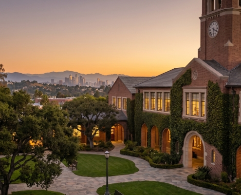 An overhead view of a historic brick private school campus in Los Angeles during golden hour, featuring ivy-covered arches, a clock tower, and manicured green lawns, with the downtown L.A. skyline and distant mountains visible on the horizon