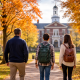 Tree-lined boarding school campus walkway leading to a classic academic building with autumn foliage and student backpacks visible from behind.