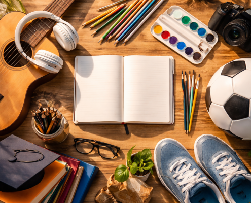 Flat lay of diverse hobbies including a guitar, soccer ball, art supplies, camera, and open notebook arranged on a wooden desk.