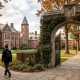 A student wearing a blazer and backpack walks along a winding stone path toward the historic red-brick buildings of an elite boarding school campus. The scene features a grand stone archway covered in ivy, lush green lawns, and vibrant autumn foliage, evoking a prestigious and scholarly atmosphere.