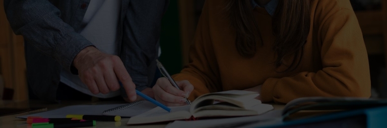 Student writing in notebook while teacher points at textbook during study session