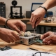 Hands collaborating on a robotics project at a wooden workbench with tools, gears, and circuit boards.