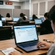 A student taking a digital exam on a laptop in a modern classroom setting, seen from behind.