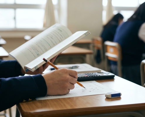 A student at a desk in a classroom, holding a book open with their left hand and a pencil in their right over a paper. Other students are visible in the background, blurred.