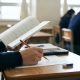 A student at a desk in a classroom, holding a book open with their left hand and a pencil in their right over a paper. Other students are visible in the background, blurred.