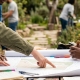 A group of people collaborating around a table outdoors, pointing at maps and project plans; no faces visible.