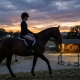 A female equestrian in professional attire riding a bay horse at dusk, with a high-end stable and sunset hills in the background.