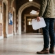 A student stands in a college hallway holding a notebook and papers.