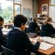 Diverse students in navy school uniforms sit at desks in a bright classroom, focused on writing in notebooks and reading textbooks.