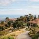 A wide landscape view of a Mediterranean-style school campus with terracotta tile roofs, nestled on a golden, grassy hill overlooking a valley and the ocean under a clear blue sky.
