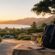 Backpack and notebook on a stone ledge overlooking a scenic coastal landscape at sunset, with mountains and trees in the background.
