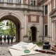 An acceptance letter with a green wax seal, a fountain pen, and leather journals resting on a stone ledge in front of a prestigious, historic school campus courtyard.