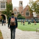 A student with a backpack walks onto a private school campus toward an athletic field and groups of students.