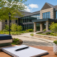 Modern school campus courtyard with a stone-and-glass building, landscaped greenery, and a wooden table holding books, an open notebook, and a small potted plant in the foreground.