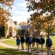 Students with backpacks walk along a pathway on a scenic, tree-lined prep school campus during autumn.