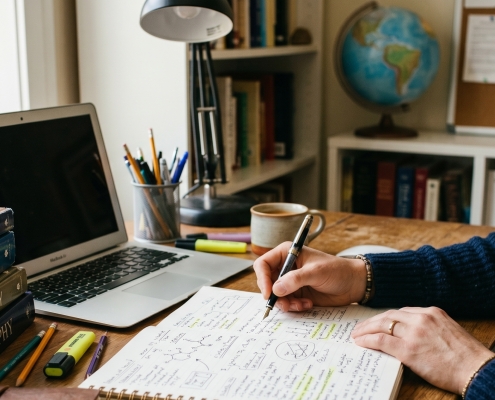 An individual writes in a notebook at a wooden desk surrounded by a laptop, a globe, and a stack of textbooks including physics, economics, and philosophy.