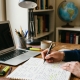 An individual writes in a notebook at a wooden desk surrounded by a laptop, a globe, and a stack of textbooks including physics, economics, and philosophy.