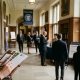 An elegant, wood-paneled hallway of an elite private school filled with parents during an open house event. In the foreground, a person holds a leather portfolio and a fountain pen, taking notes on an admissions brochure. The background shows attendees walking through the hall, framed by large leaded-glass windows and oil portraits. All faces are obscured or turned away, and any text is illegible.