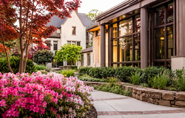 Modern private school campus courtyard with landscaped garden, blooming flowers, and contemporary academic buildings in soft daylight.
