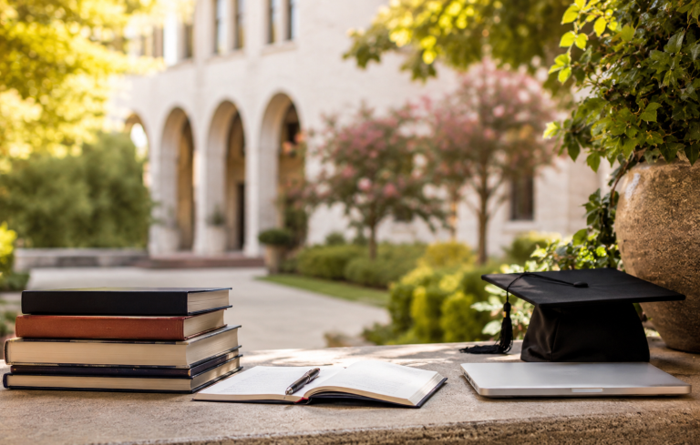 Books, notebook, and graduation cap on a stone ledge with a blurred academic campus courtyard and arched building in the background.