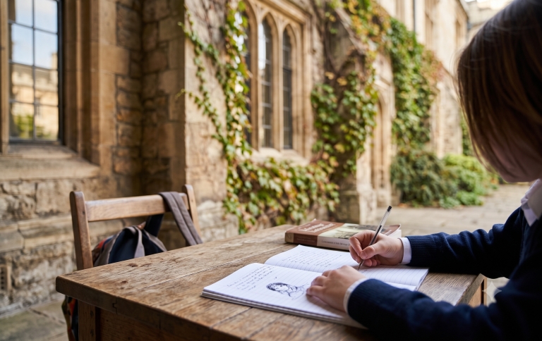 A student in uniform writes in a notebook at an outdoor wooden desk.